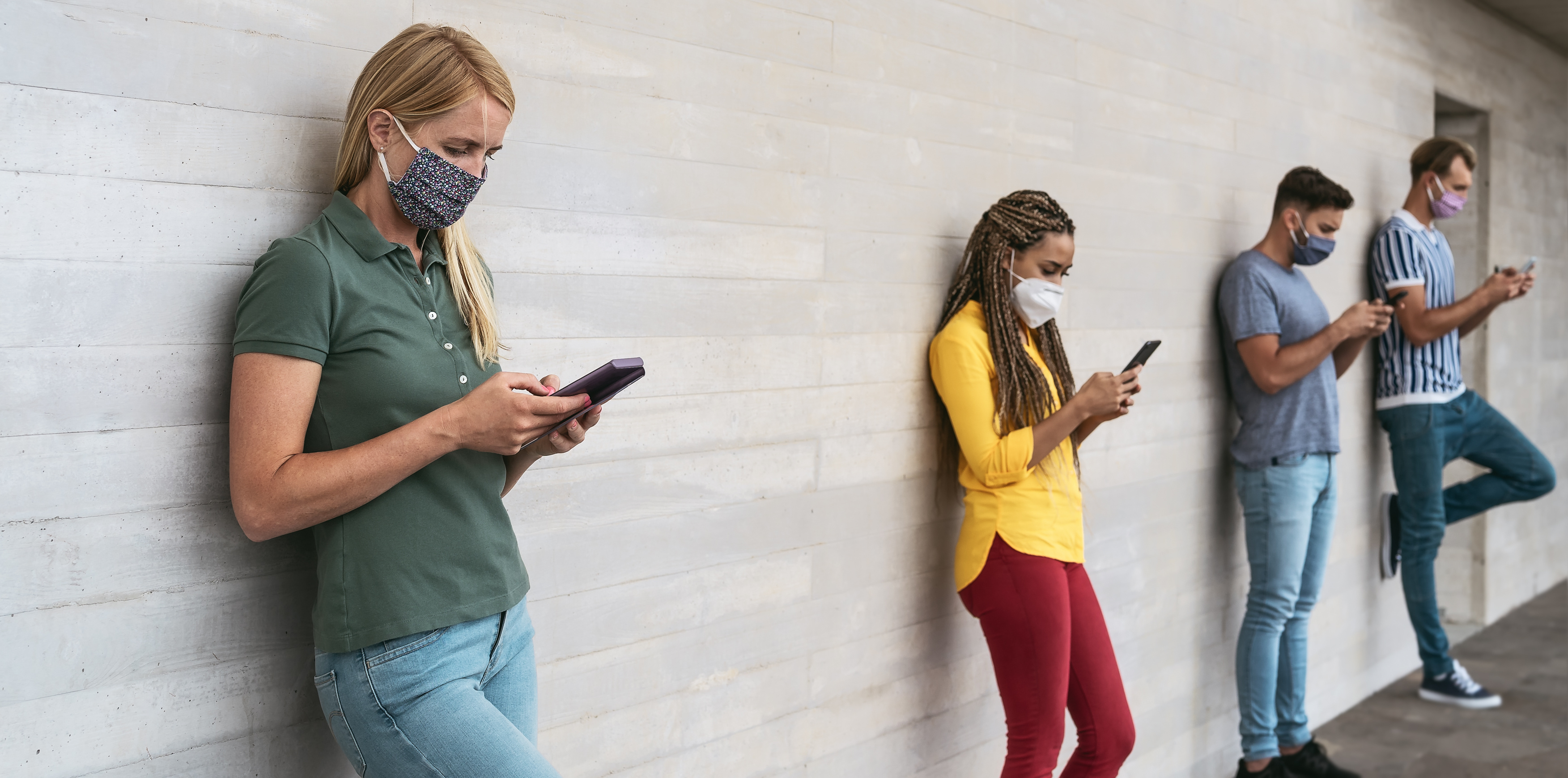 Vier Personen mit Masken und Smartphones lehnen an der Wand und betrachten die Smartphones.